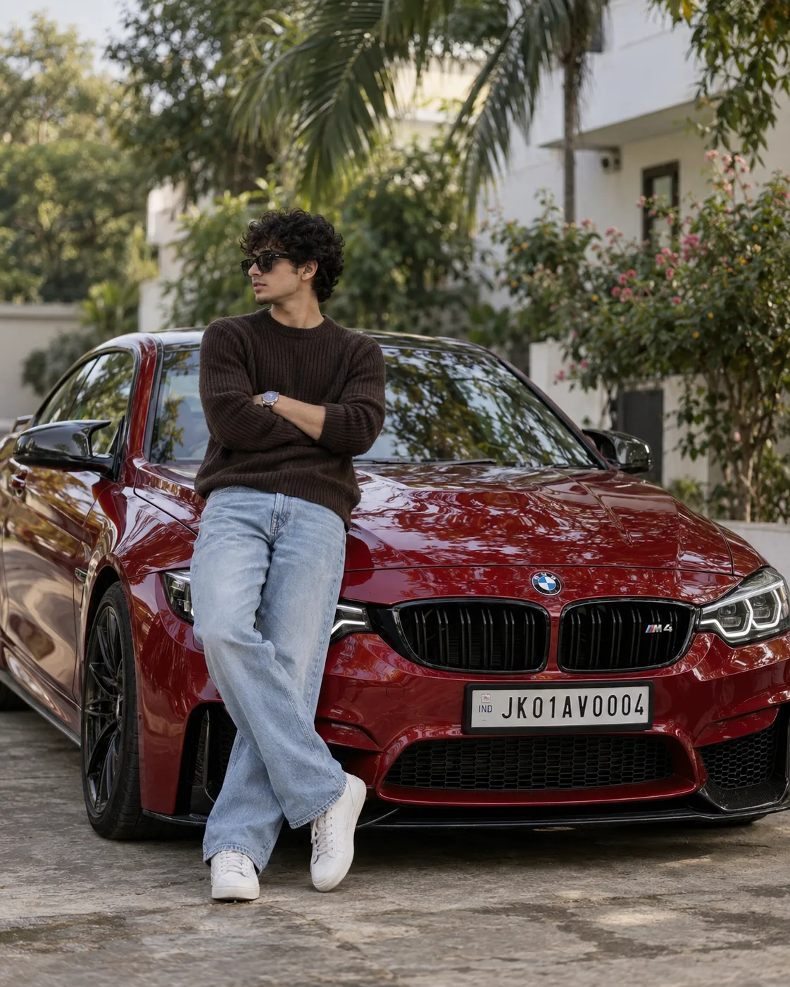 A stylish young man with curly hair, wearing black sunglasses, leaning casually against a shiny red BMW M4 in an outdoor residential