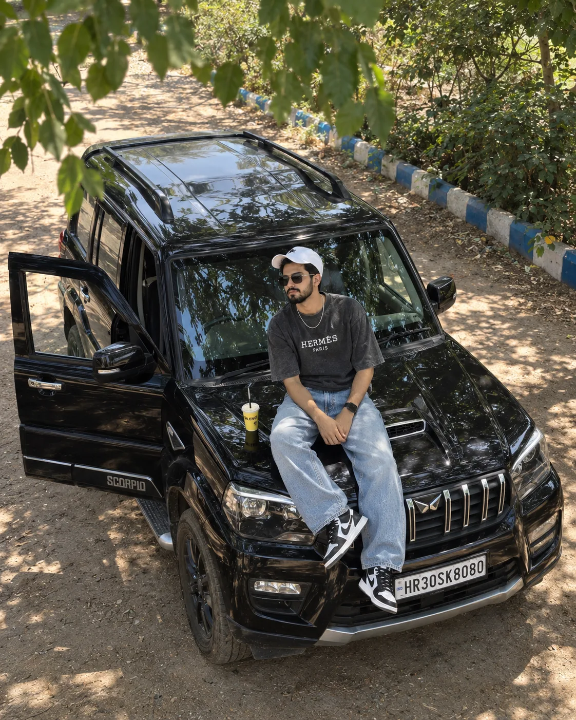 A high-angle photo of a stylish young South Asian man in his 20s sitting casually on the hood of a glossy black Mahindra Scorpio Classic