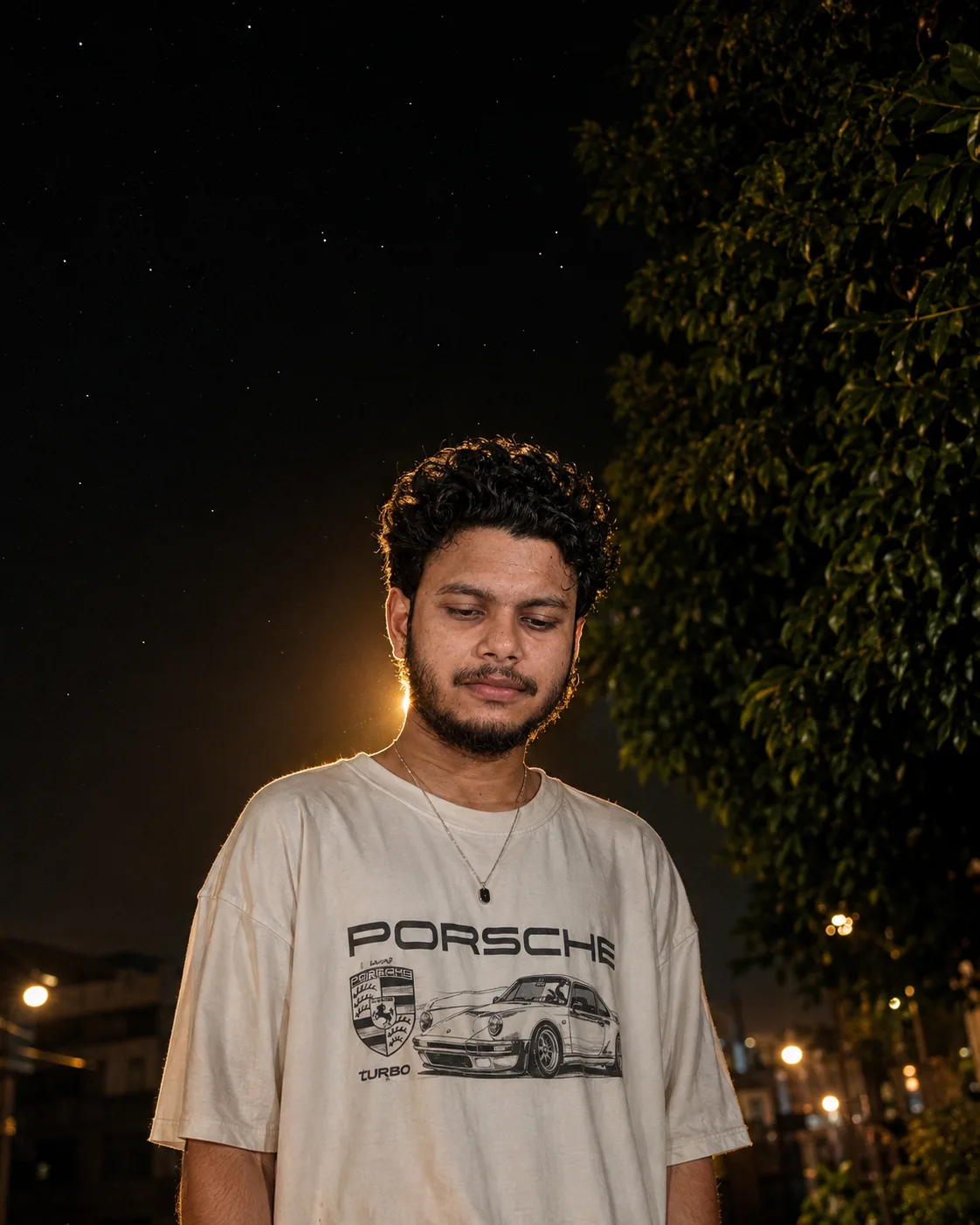 A realistic night-time portrait of a young Indian man in his early 20s standing outdoors under a dark starry sky.