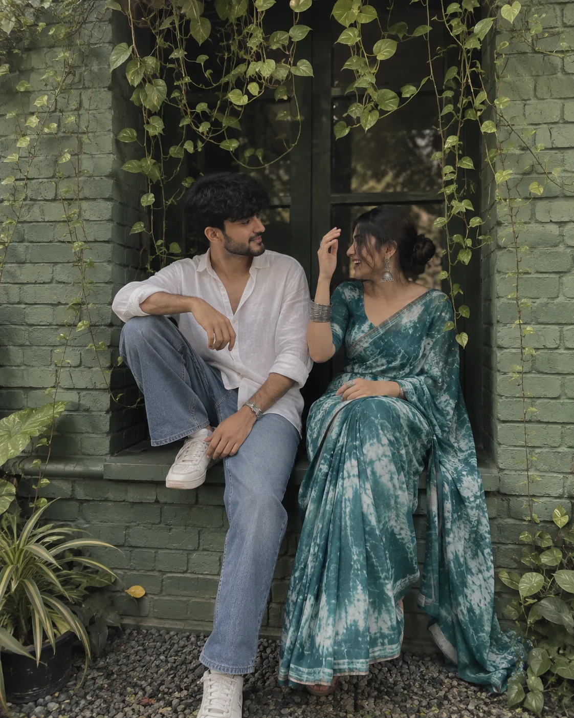 INDIAN COUPLE SITTING ON A GREEN-PAINTED BRICK WINDOW WOMAN LEDGE, OVERGROWN CREEPER VINES HANGING ABOVE.