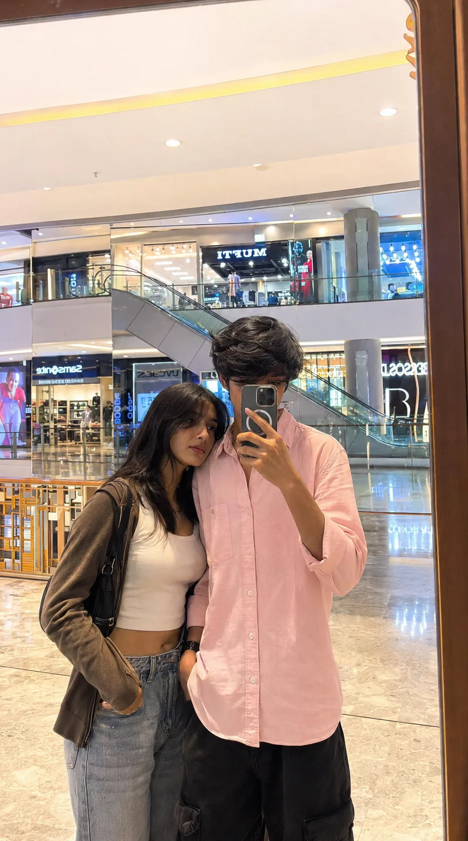 create an ultra-realistic mirror selfie of a teen young couple inside a modern shopping mall, captured like a genuine candid iphone photo.