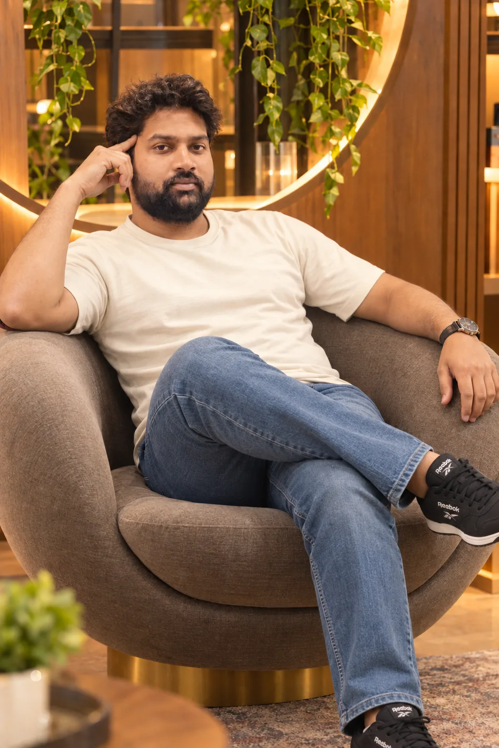 A photorealistic, medium-shot portrait of the young man seen in image_1.png, with his characteristic curly dark hair and beard, sitting in