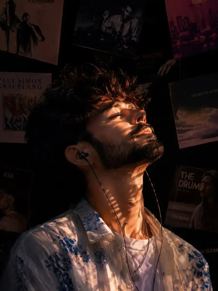 Ultra-realistic cinematic portrait of a young South Asian man sitting indoors in a dark, intimate music-themed room, captured in a natural
