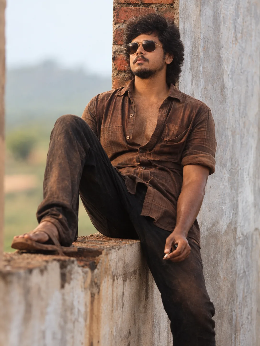 Create an ultra-realistic cinematic vertical portrait of a rugged young South Asian man sitting casually on a weathered concrete ledge