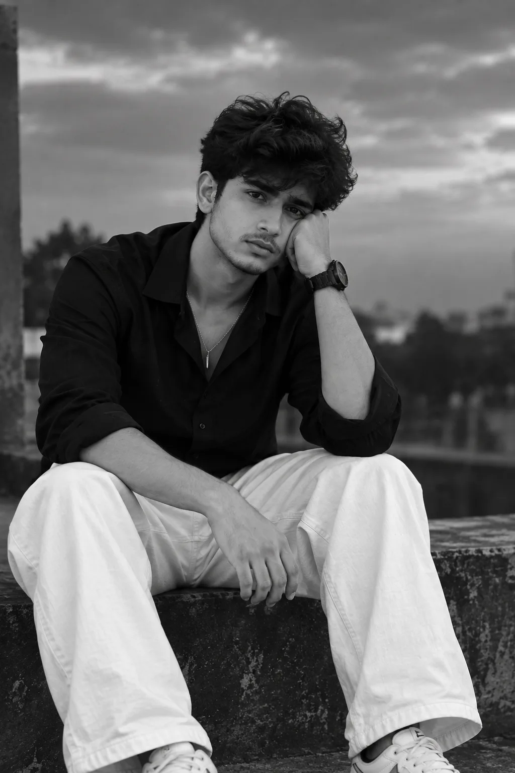 A handsome young South Asian man sitting alone on a rooftop ledge in a sad, emotional mood, **face clearly visible from the front**,