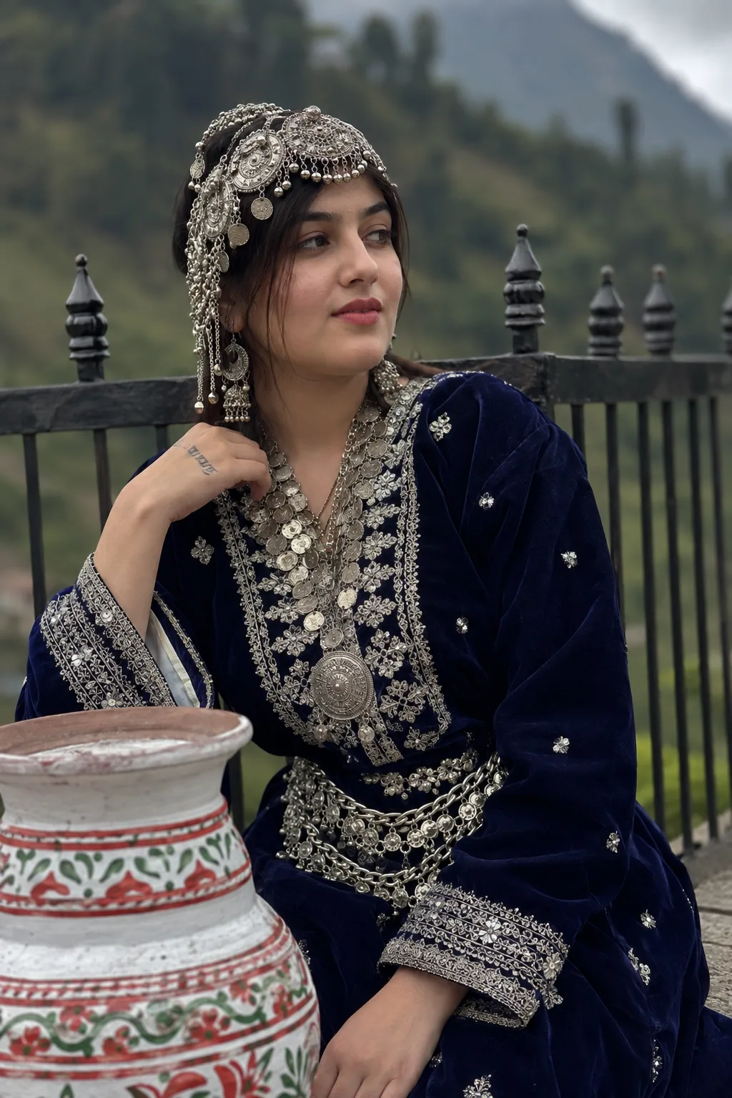 A young South Asian woman in traditional Himachali attire, medium portrait shot, leaning against a black wroght-iron fence with decoratiy