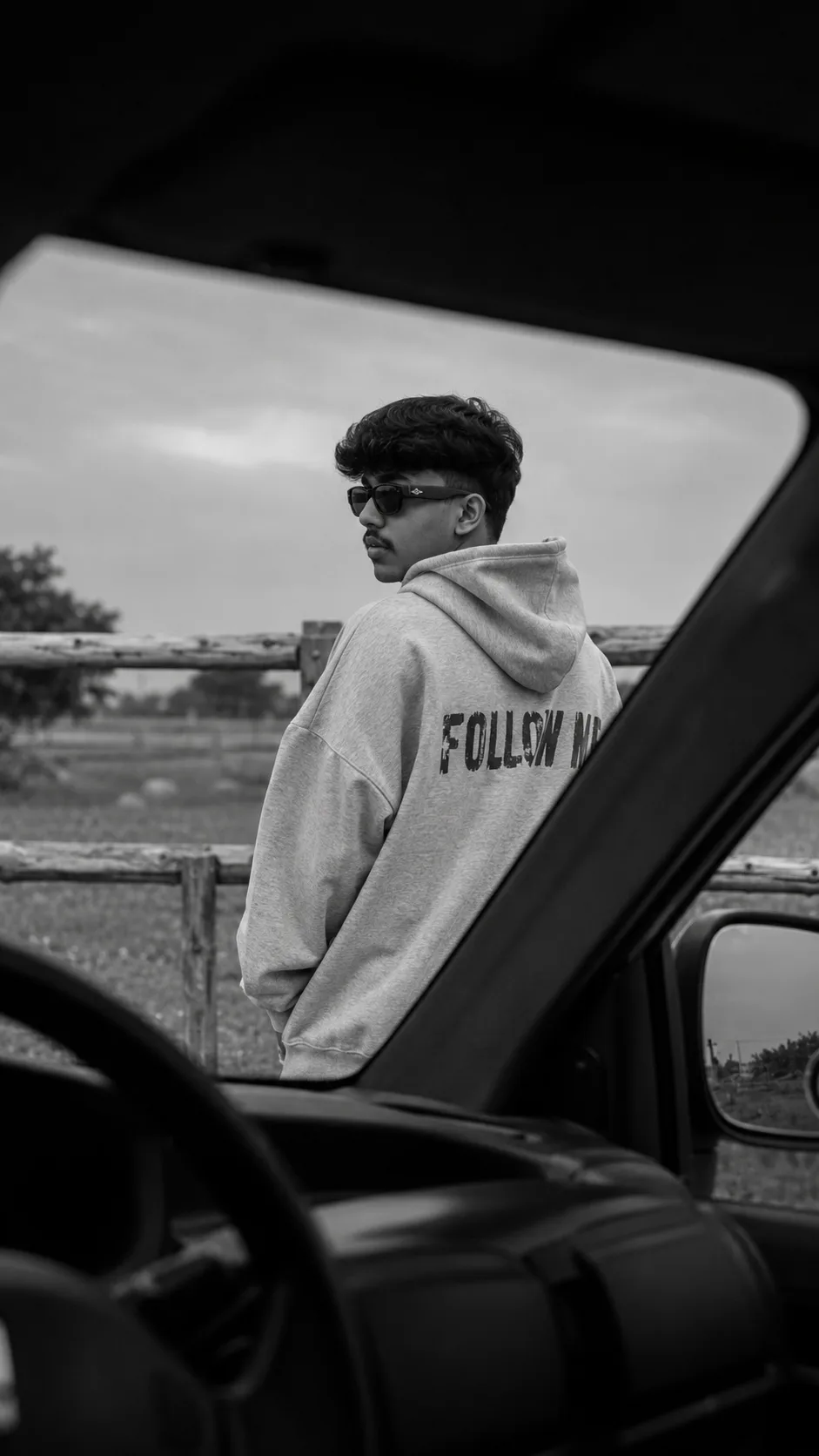 Ultra-realistic black and white cinematic photo of me standing outside near a wooden fence in an open rural area, viewed from inside a