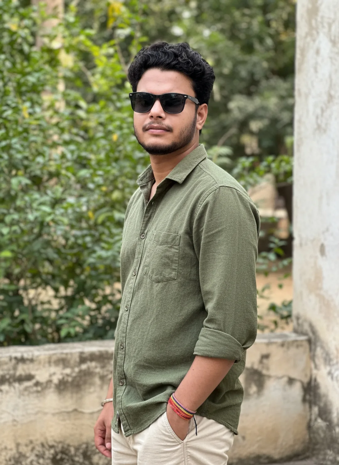 A young South Asian man with short curly black hair, light beard and mustache, wearing black rectangular sunglasses, standing outdoors with