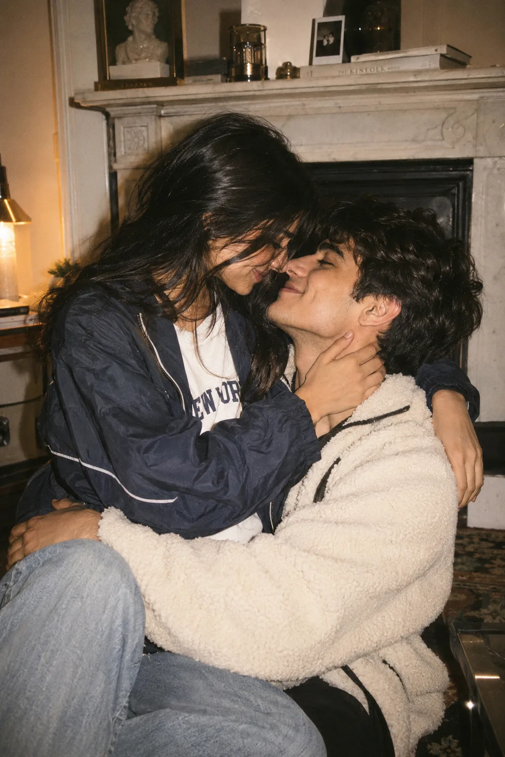 A candid, intimate lifestyle photograph of a young couple sitting closely together indoors in front of a white marble fireplace.