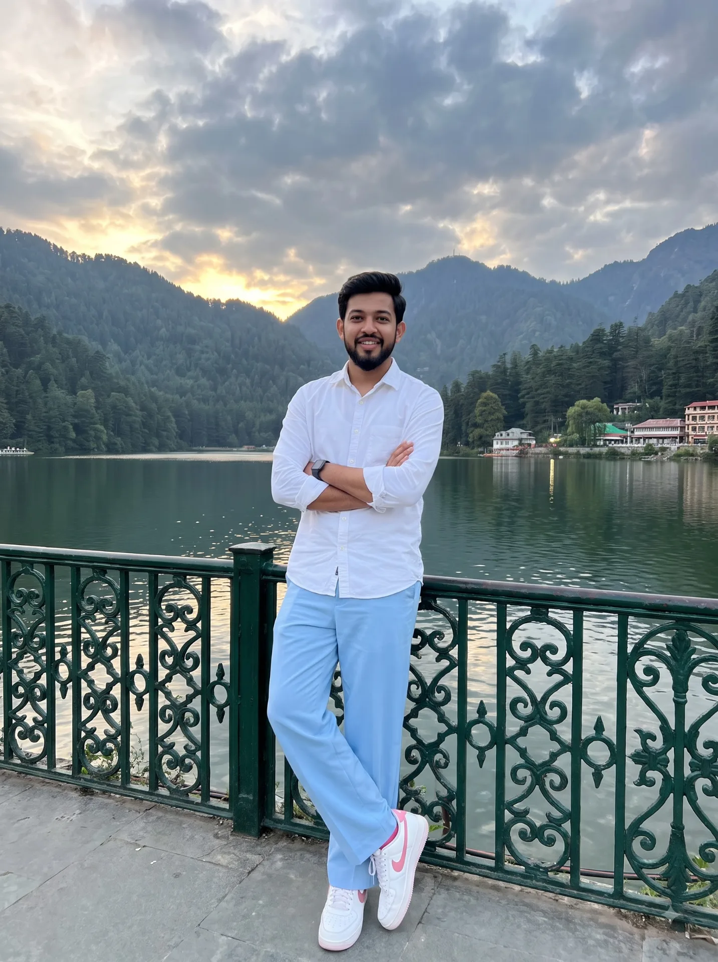 A REALISTIC TRAVEL PORTRAIT OF A YOUNG INDIAN MAN STANDING IN FRONT OF A SCENIC MOUNTAIN LAKE WITH HIS ARMS CROSSED CONFIDENTLY.
