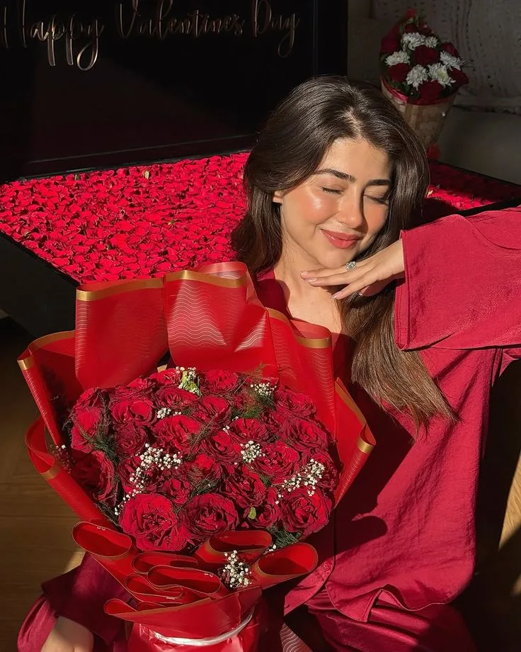 A romantic Valentine’s Day portrait of a young woman sitting beside a bed covered with scattered red rose petals, holding an enormous