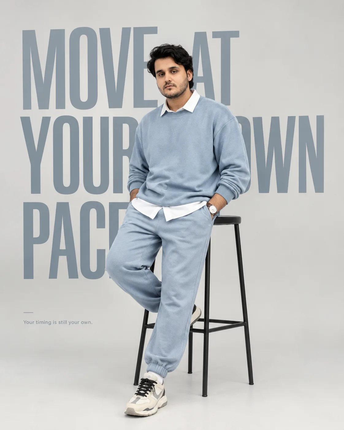 Premium full-body studio fashion photography of a young Indian male model with styled dark textured hair, leaning casually against a tall,