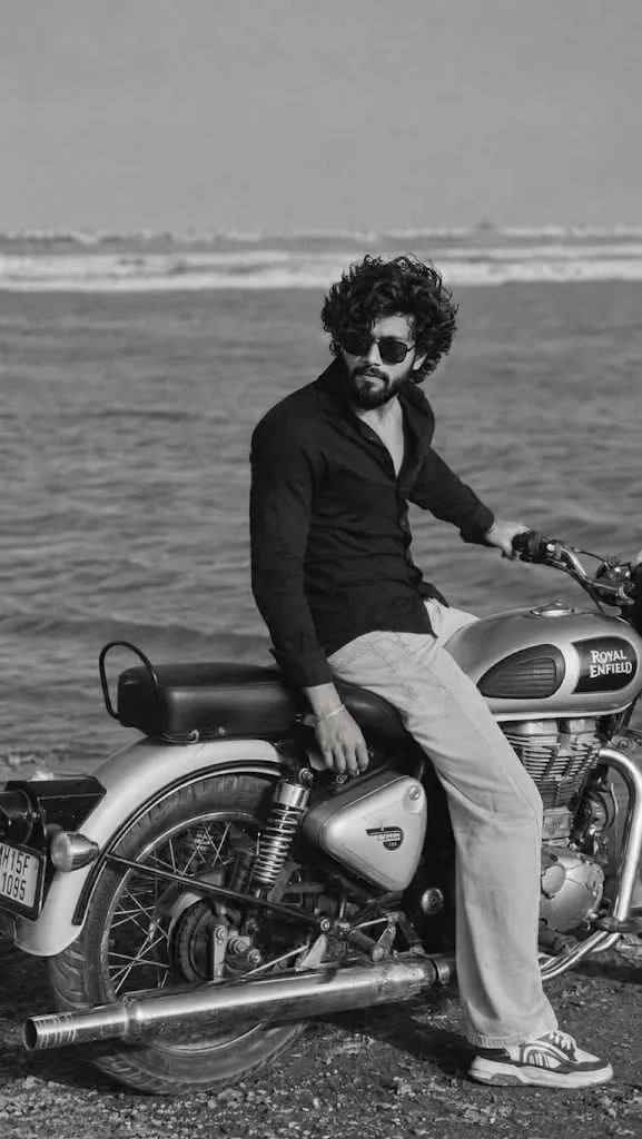 A cinematic black-and-white fashion photograph of a young man sitting sideways on a classic Royal Enfield motorcycle beside the ocean.