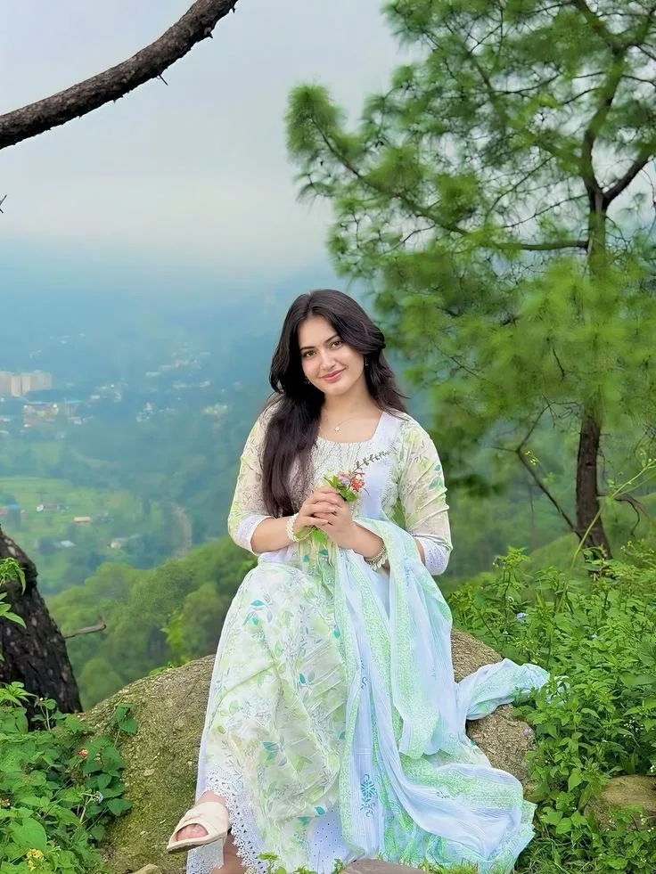 Create a photorealistic vertical 9:16 portrait of a young Indian woman sitting gracefully on a natural rock at a beautiful mountain