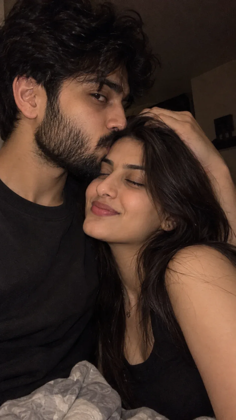 Photorealistic romantic close-up selfie of a young couple cuddling cheek-to-cheek in a dimly lit room.
