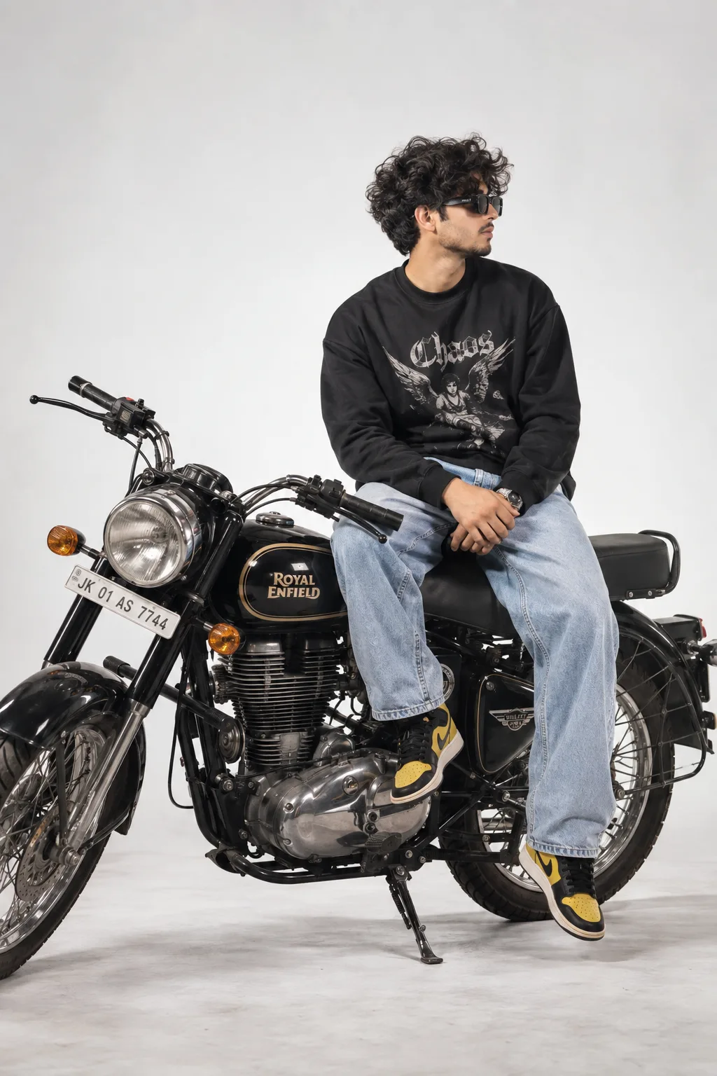 A stylish young man with curly hair, wearing black sunglasses, sitting on a black Royal Enfield-style vintage motorcycle in a minimalist