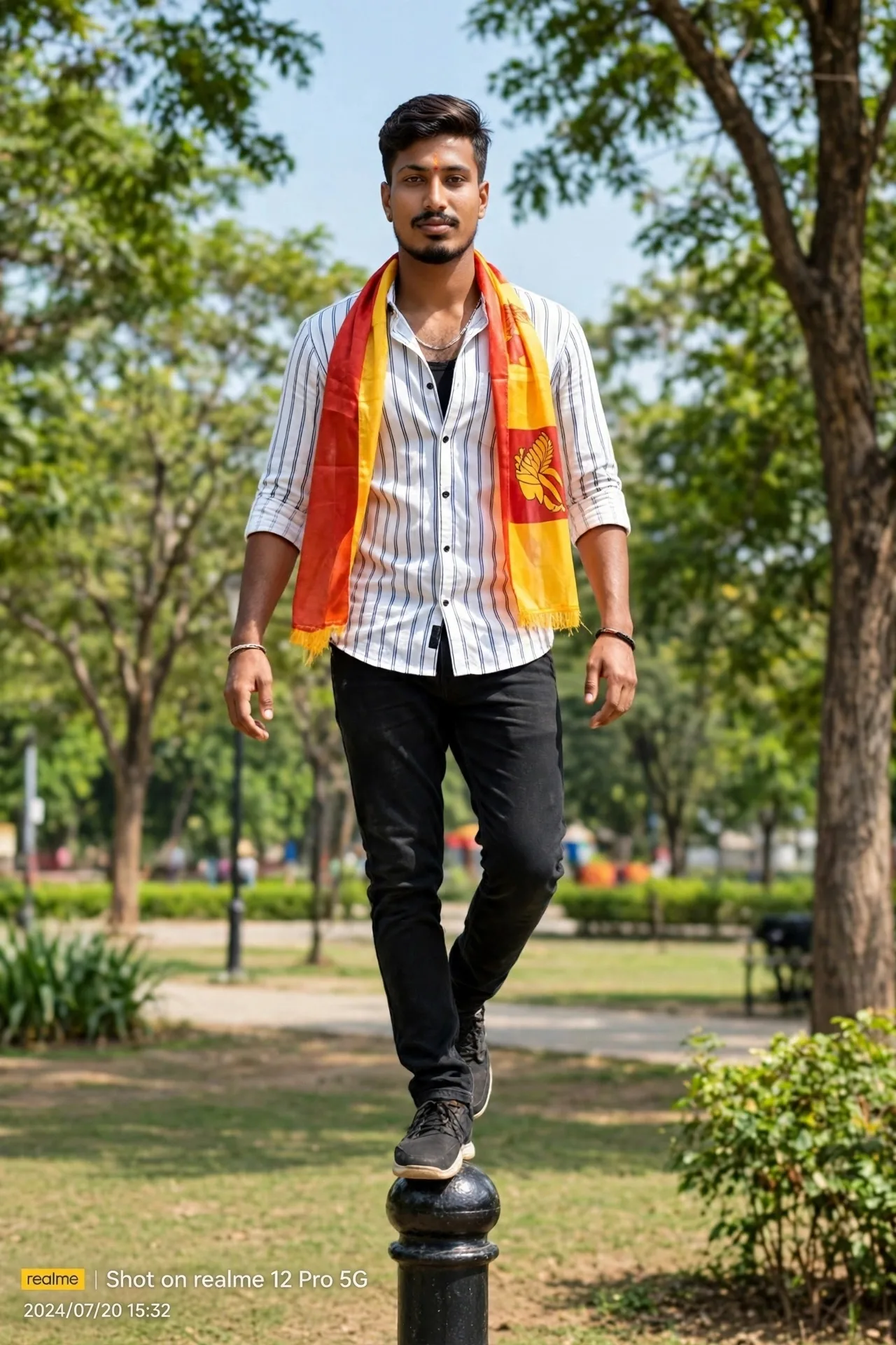 A young Indian man balancing on a black bollard in an outdoor park, wearing white striped shirt, black jeans, red and yellow Karnataka flag