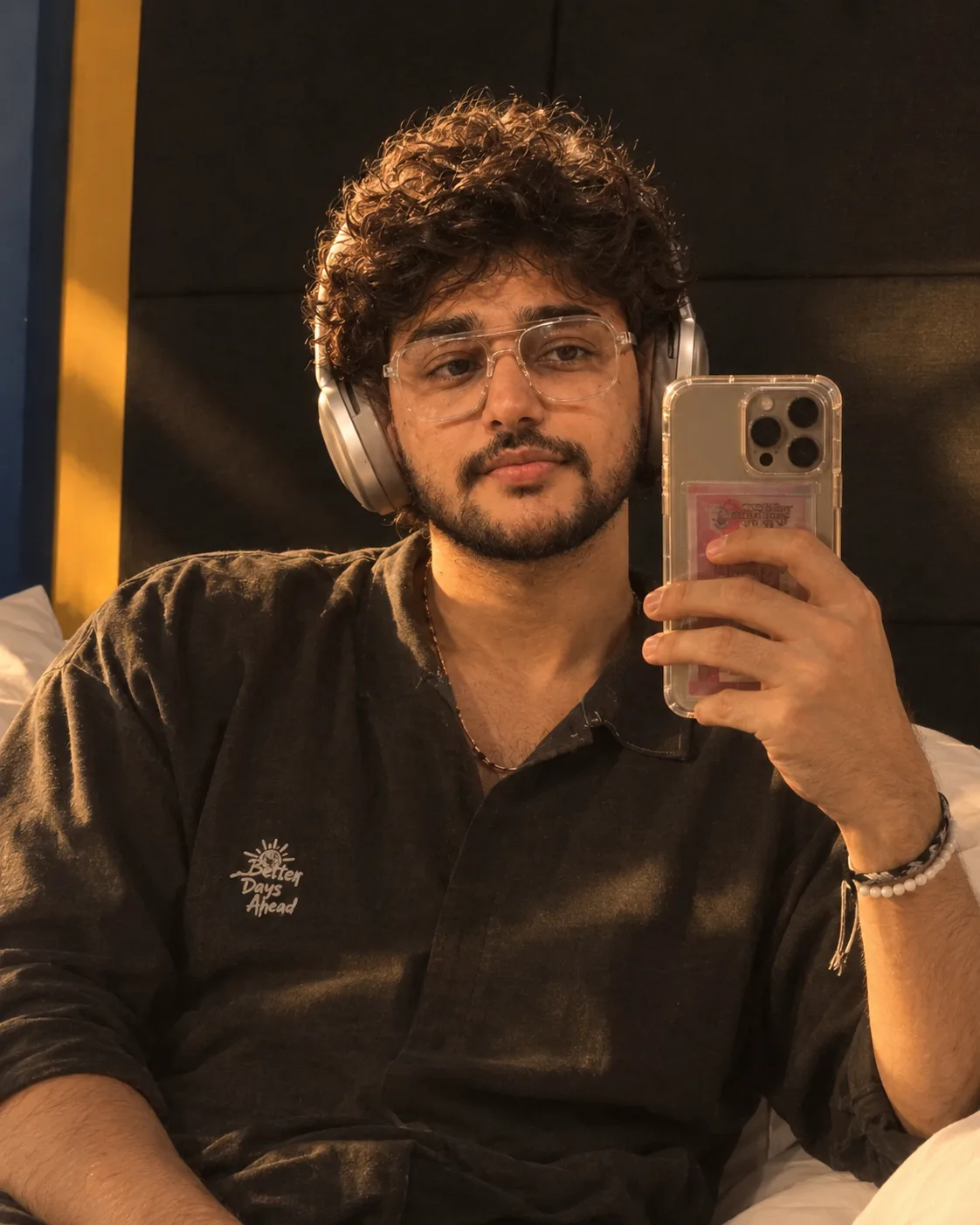 "A warm, golden-hour mirror selfie of a young man with thick, soft, tousled dark brown curly hair, smiling gently as he looks at his