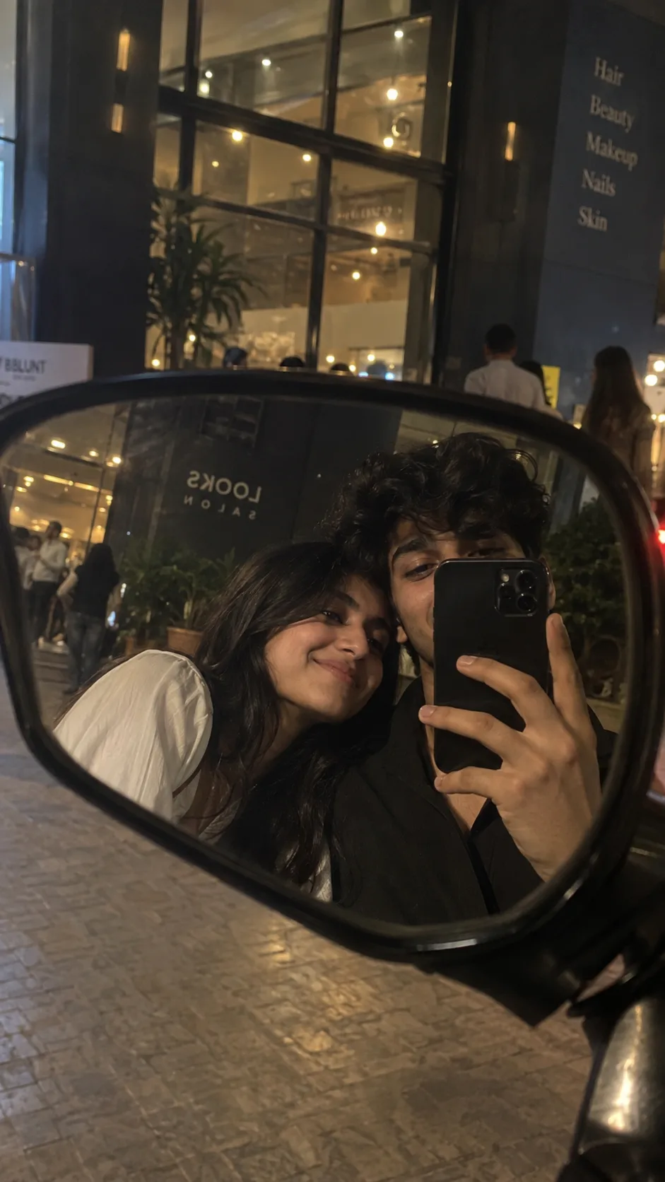 Photorealistic candid nighttime smartphone photo of a young couple taking a close selfie through a car/bike side mirror.