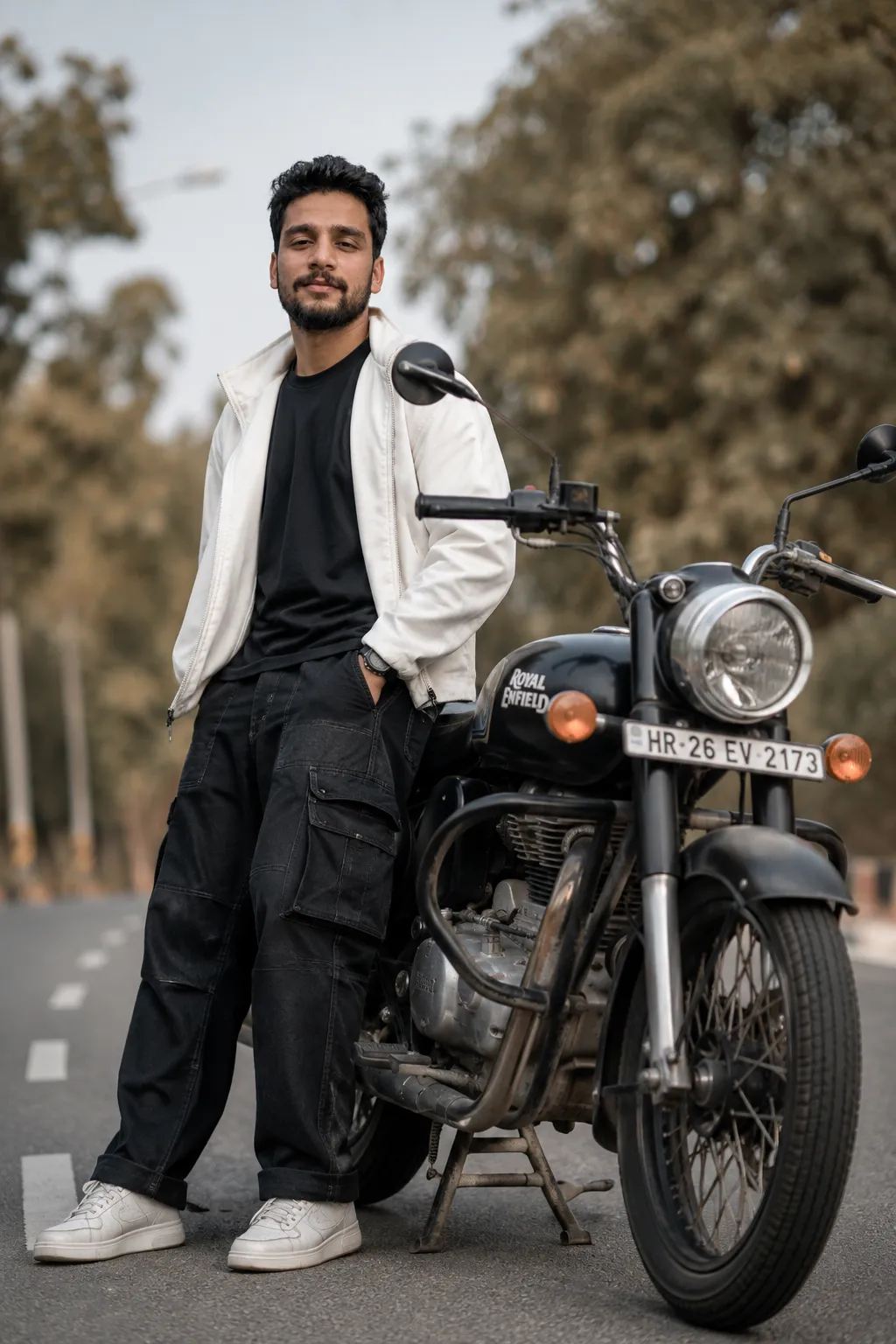 A young man standing confidently beside a Royal Enfield Bullet 350, wear-ing black baggy jeans with visible side pockets, a solid t-shirt,