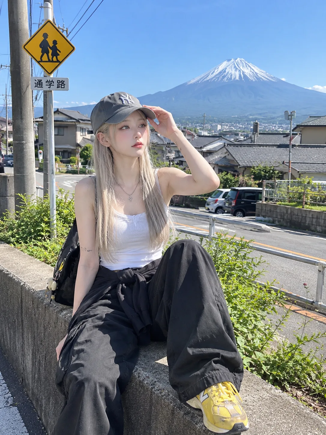 A realistic photo of a young woman sitting casually on a concrete ledge by the roadside on a bright, sunny day, with Mount Fuji clearly