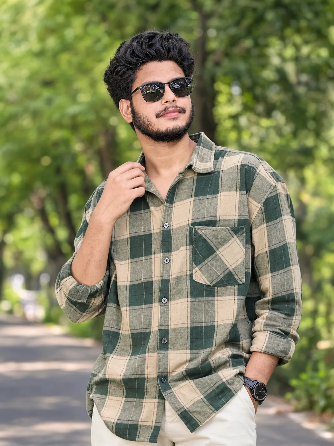 Prompt: A young man with curly black hair and a short beard, wearing black rectangular sunglasses, standing outdoors on a tree-lined paved
