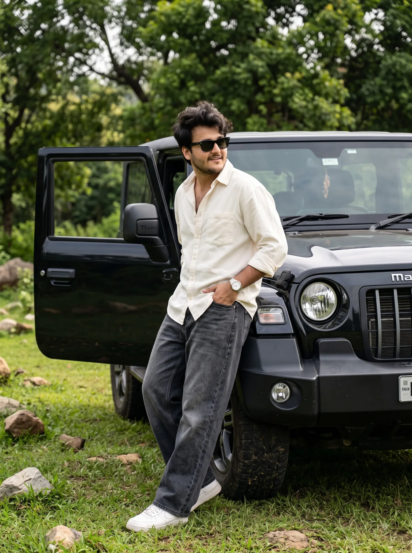 A casual lifestyle portrait of a young man leaning against the front of a black Mahindra Thar SUV in a natural outdoor setting.