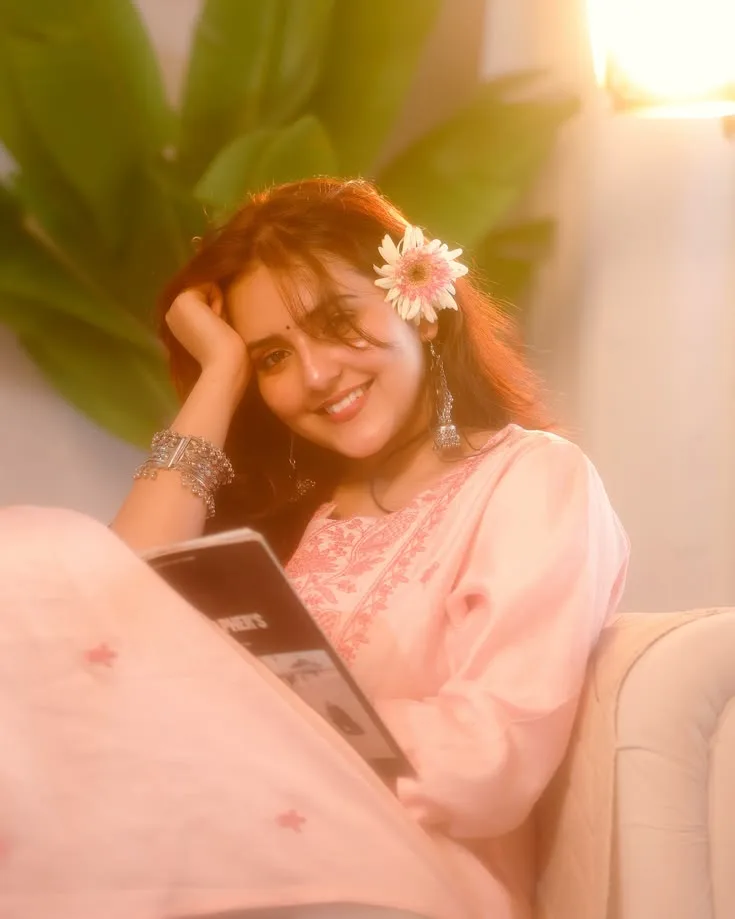 Create a dreamy, warm-toned photorealistic portrait of a young woman relaxing comfortably on a sofa, wearing a soft pastel pink traditional