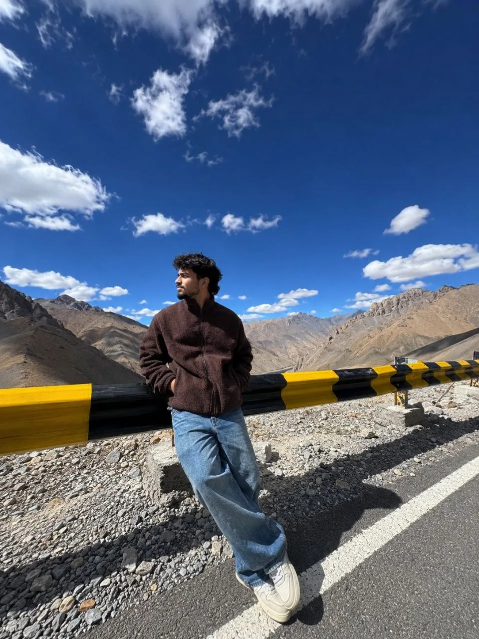 Ultra-realistic iPhone 15 Pro ultra-wide lens photo of a young man leaning casually against a yellow-and-black mountain road guardrail on a
