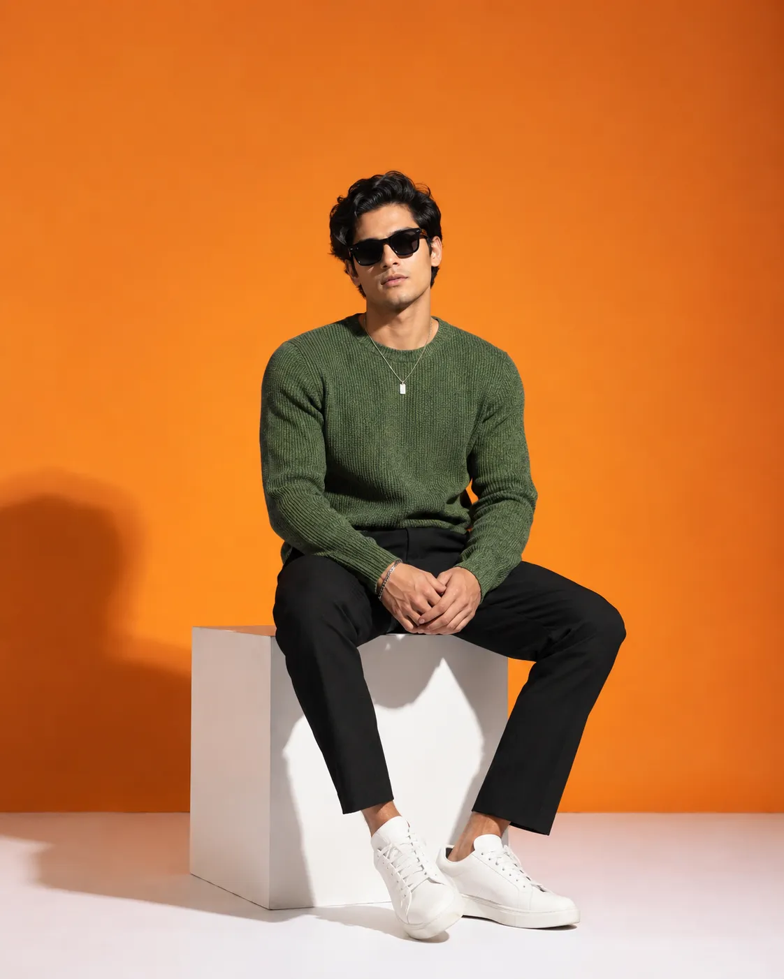 A stylish male model sitting on a white cube in a minimalist studio with a vibrant orange background.