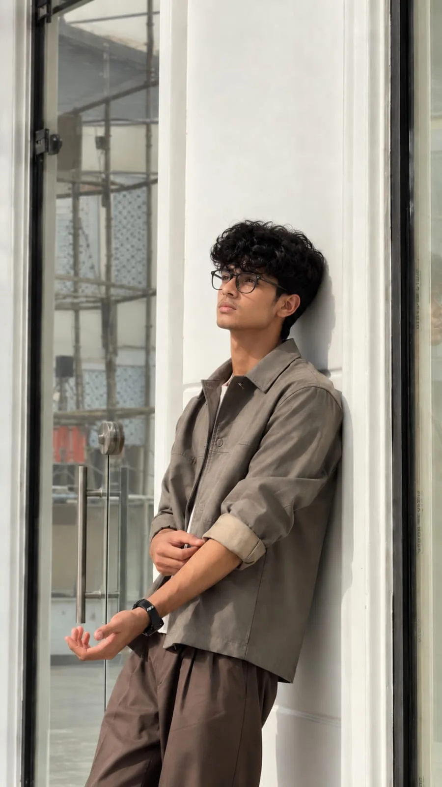 Ultra-realistic full-body portrait of a young man leaning casually against a clean white architectural wall beside a large glass door.