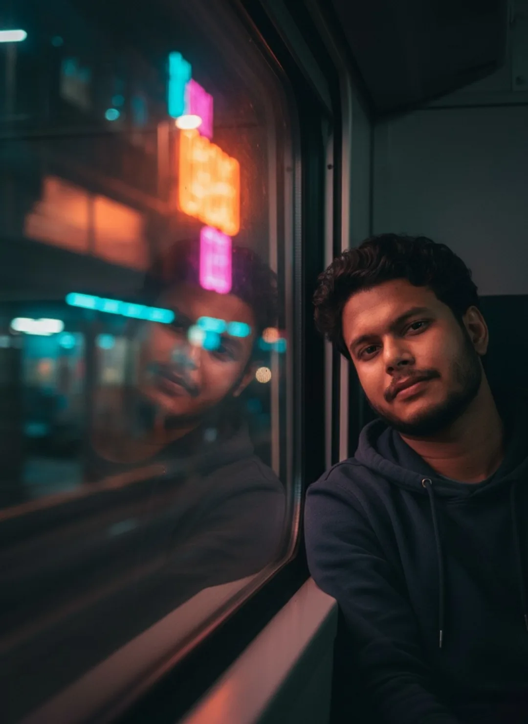 A young South Asian man with curly dark hair and light beard, wearing a dark navy hoodie, sitting by a train window at night, leaning his
