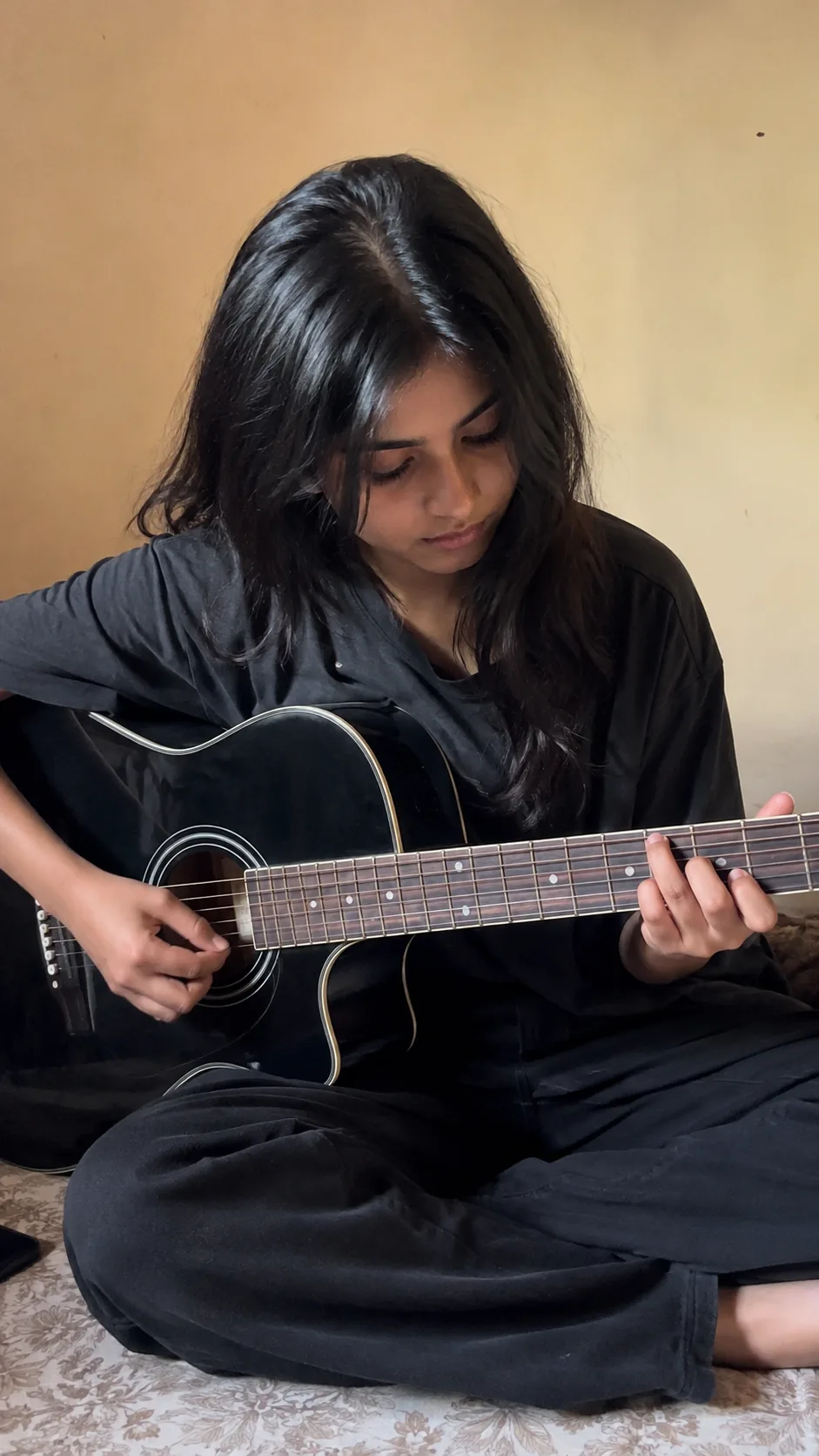 Ultra-realistic smartphone portrait of a young woman sitting cross-legged indoors while playing a black acoustic guitar.