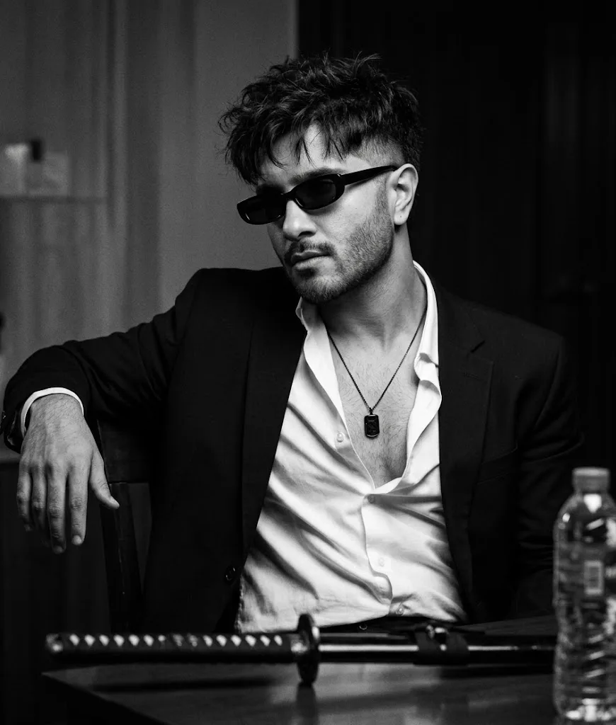 Ultra-realistic cinematic black-and-white portrait of a stylish young man sitting casually at a table in a dark moody room.