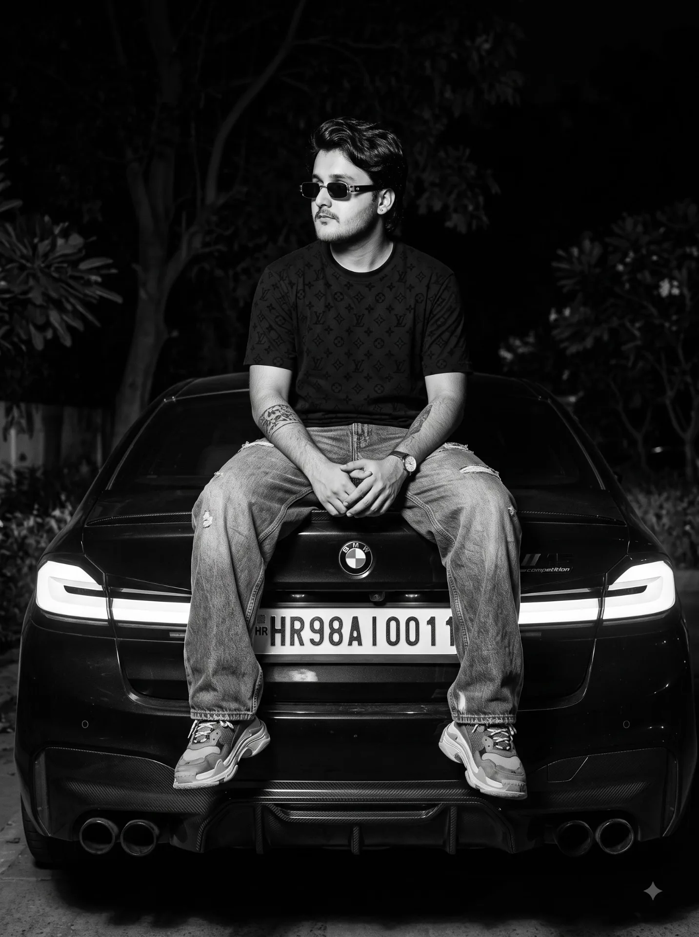 Cinematic black and white night portrait of a young South Asian man in his early 20s with stylish black hair and a light beard, sitting on