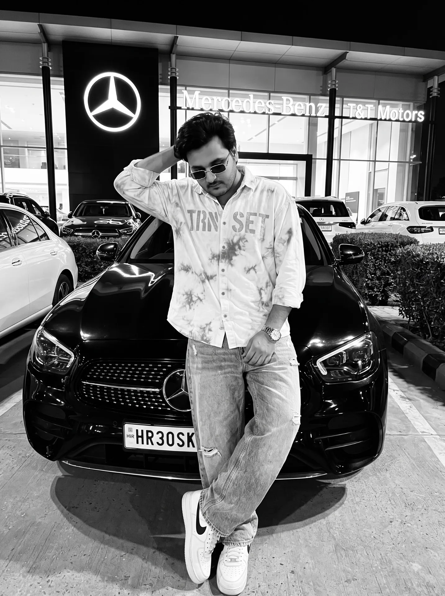 Black and white night flash photo of a young Indian man with stylish black hair and light beard, wearing an oversized white acid-wash