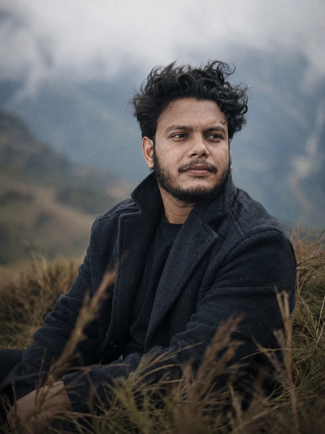A realistic portrait of a young South Asian man in his mid-20s sitting outdoors in a grassy hillside.