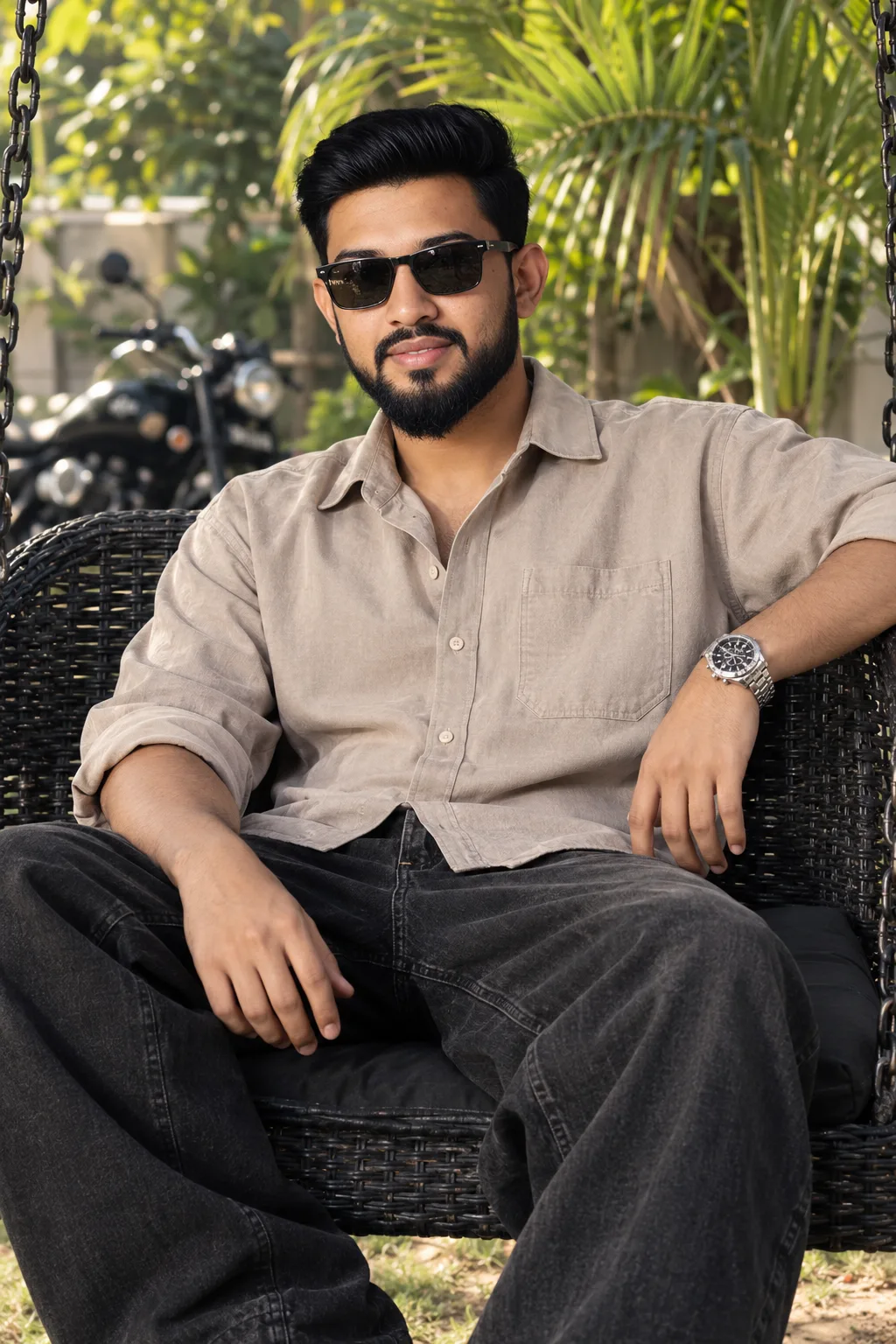 Same exact man sitting on black wicker garden swing, same face, same hairstyle, same black sunglasses, same garden background with palm