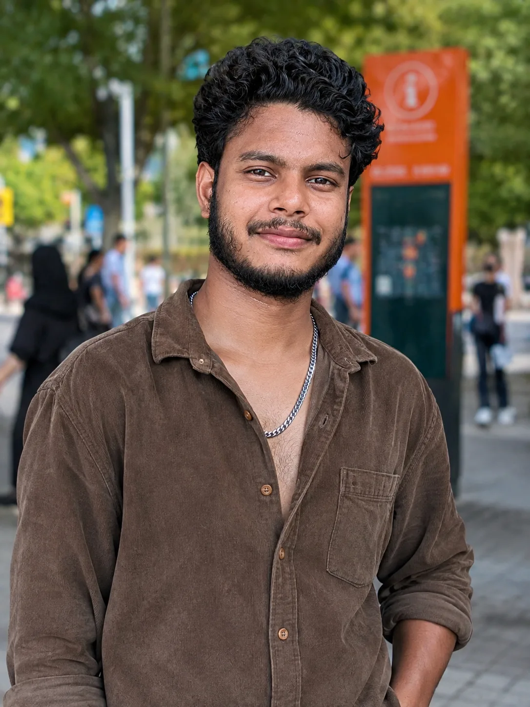 Prompt: "A young man with tight curly dark hair and a well-groomed short beard, smiling warmly at the camera, wearing an unbuttoned brown