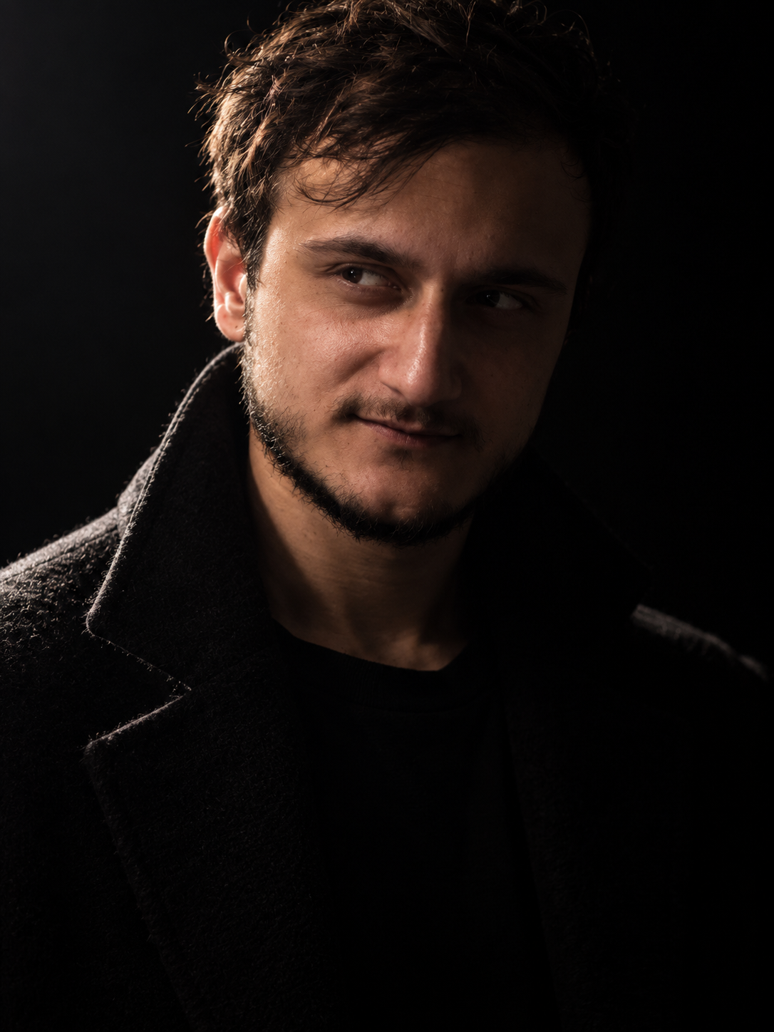 A mysterious man with messy textured hair emerging from darkness, dramatic side lighting, black background, intense expression, moody