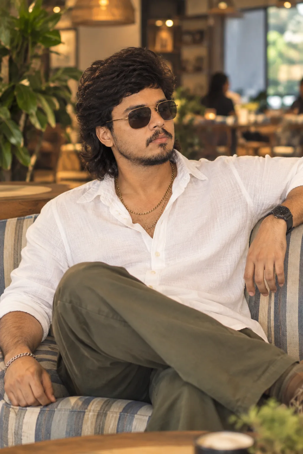 Relaxed café portrait of a young man seated on a striped sofa, leaning back with left arm resting on the backrest and right hand relaxed on