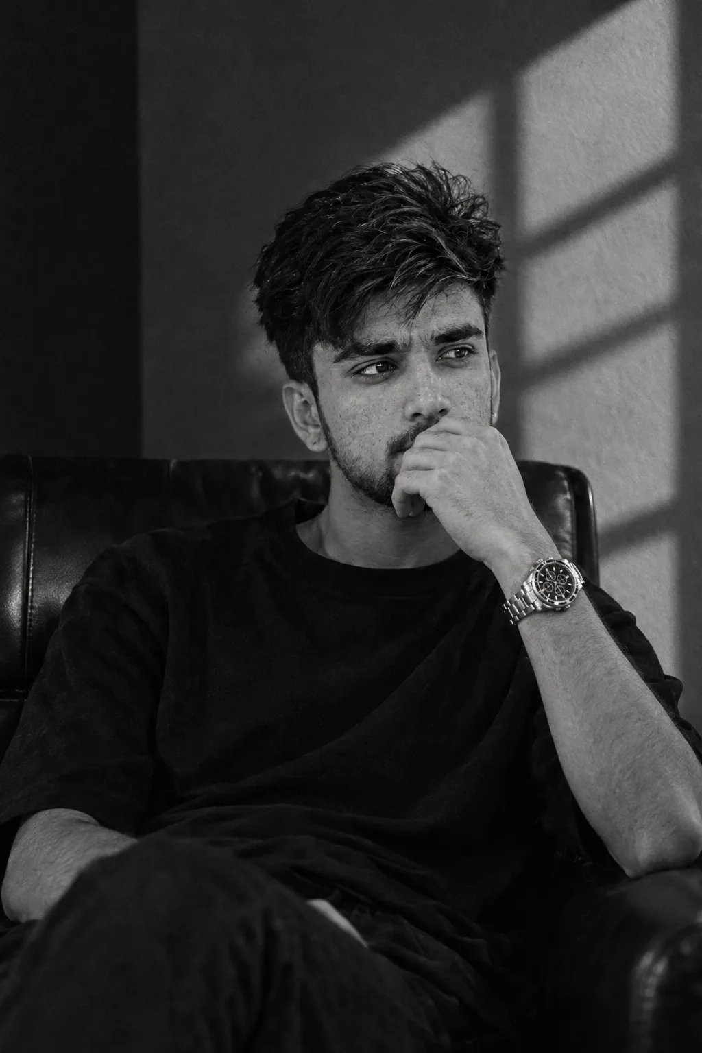 A black and white cinematic portrait of a young South Asian man with messy textured hair and light stubble, wearing an oversized plain