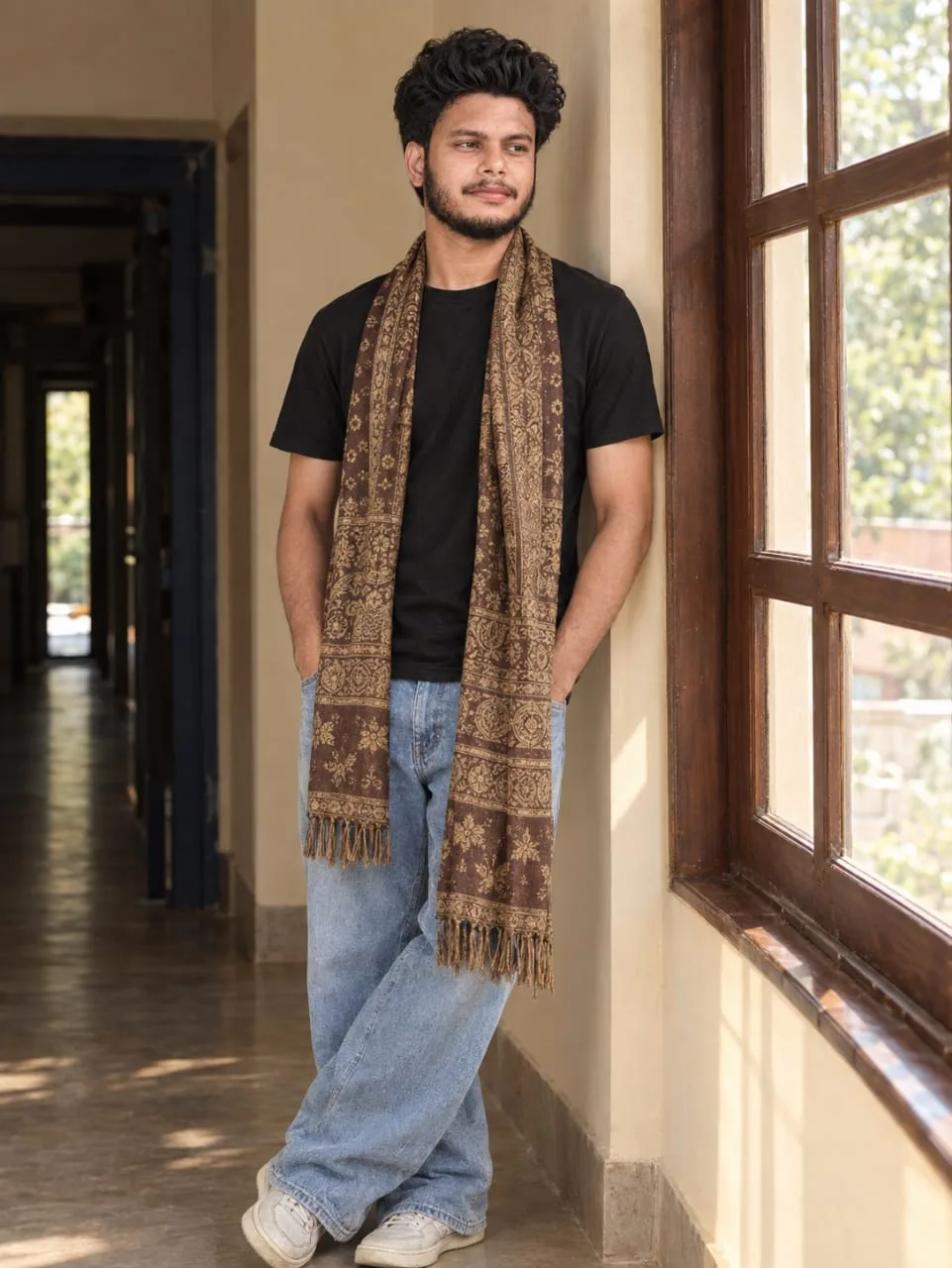 A young South Asian man with curly black hair and a short beard, leaning casually against a cream-colored wall near a large wooden-framed