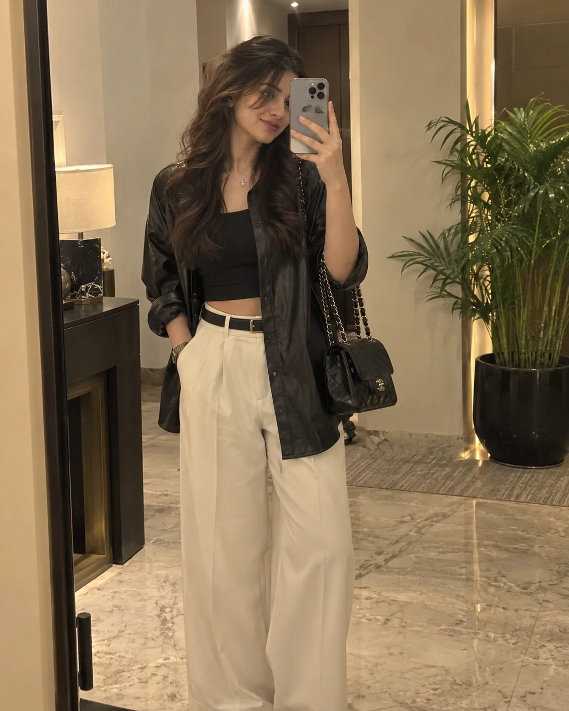 Ultra-realistic mirror selfie of a young woman in a black crop top, oversized black leather shirt, cream wide-leg trousers, and black belt,