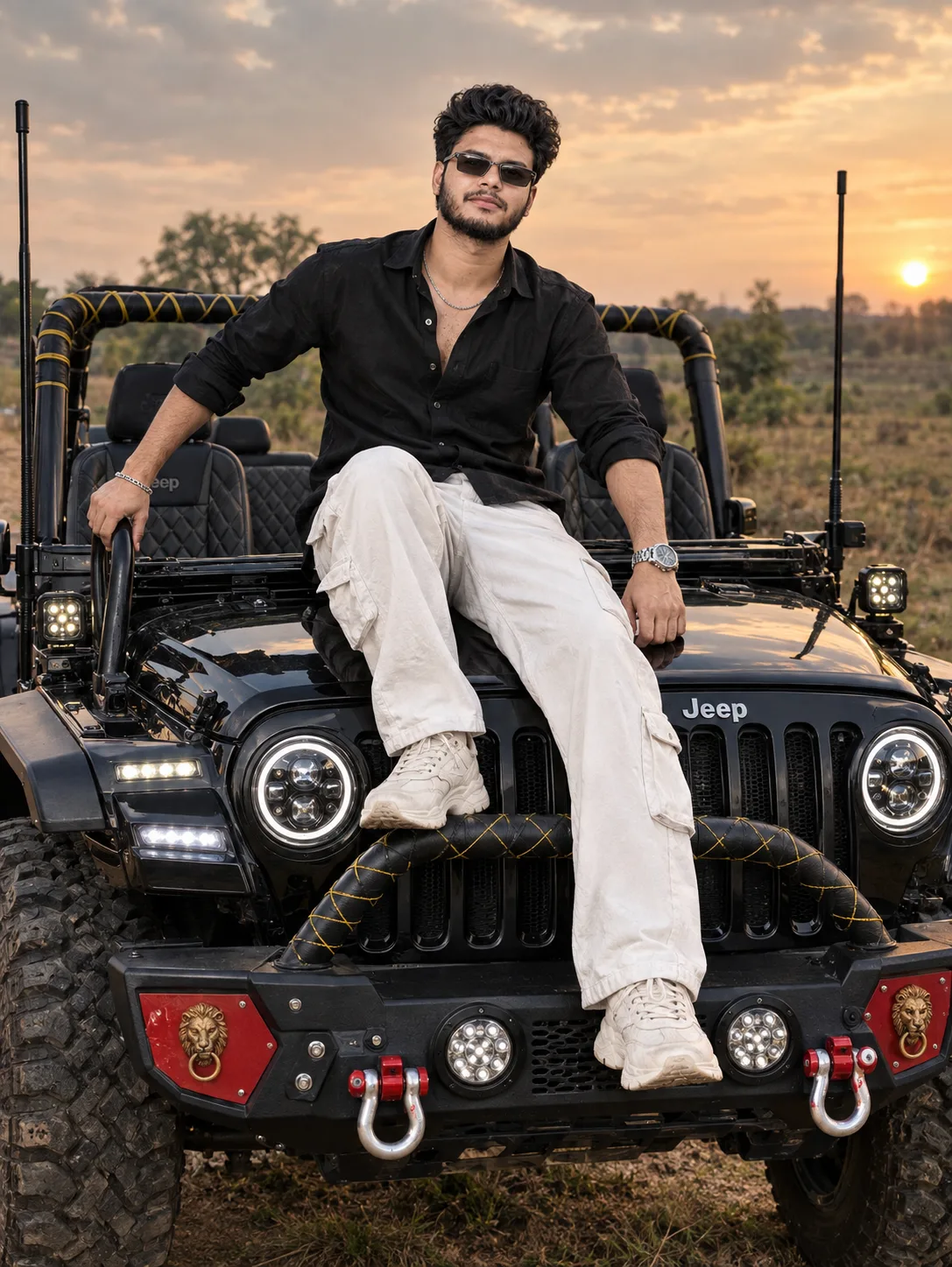 A confident young Indian man with curly dark hair, full beard, and black sunglasses sits casually on the hood of a heavily modified black