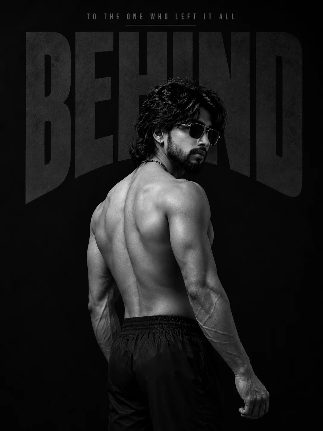A monochrome studio portrait of an athletic man viewed from a rear three-quarter angle against a dark backdrop.