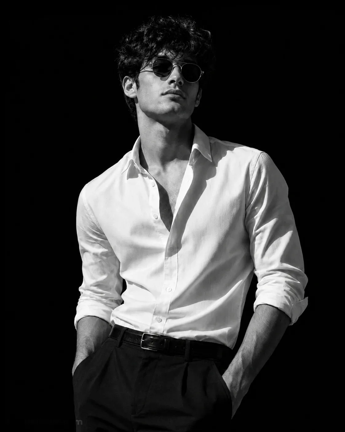 Dramatic black-and-white studio portrait of a man, wearing round vintage sunglasses.