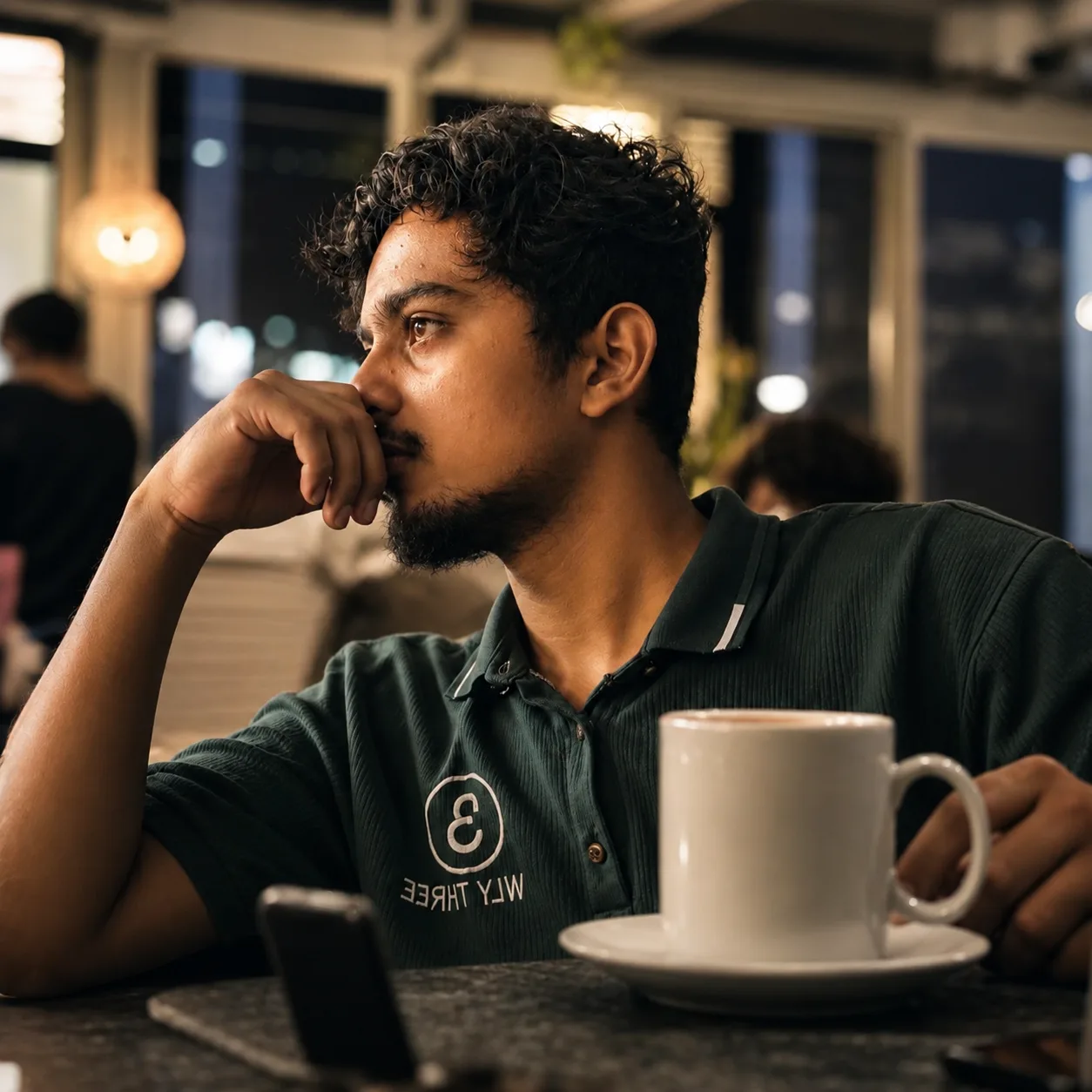 A cinematic close-up photo of a young South Asian man with short curly black hair and a light beard/mustache, sitting at a dark marble