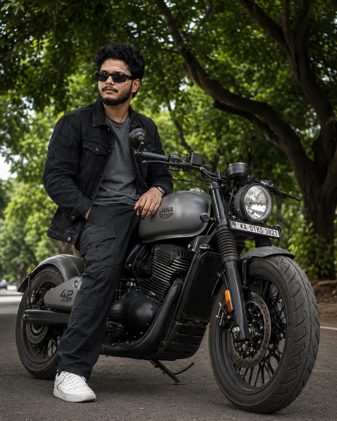 Ultra-realistic full-body shot of a stylish young man sitting confidently on a matte grey Jawa 42 bobber motorcycle under lush green trees.