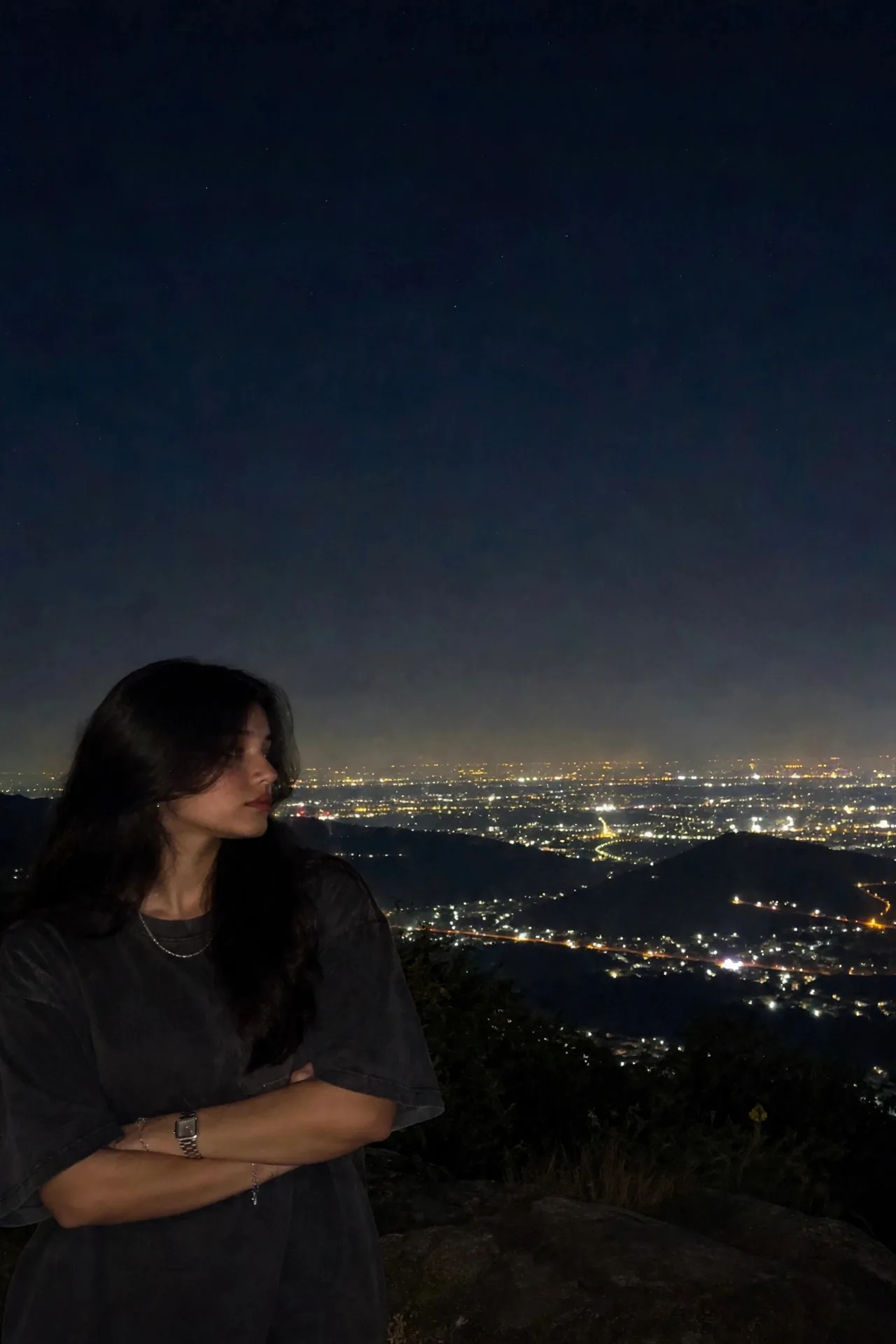 A young woman stands on a plateau at night with a vast sparkling city lights backdrop, positioned in the bottom-left of the frame while the